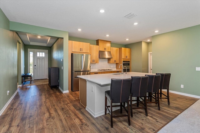 kitchen with a center island with sink, a kitchen breakfast bar, light brown cabinetry, dark hardwood / wood-style flooring, and stainless steel refrigerator