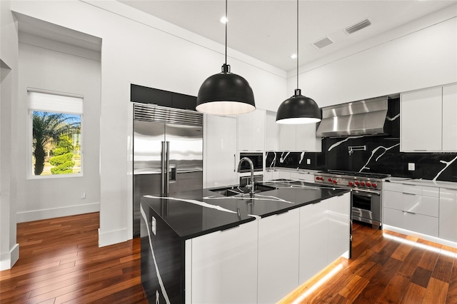 kitchen with white cabinets, hanging light fixtures, high end appliances, wall chimney range hood, and a center island with sink