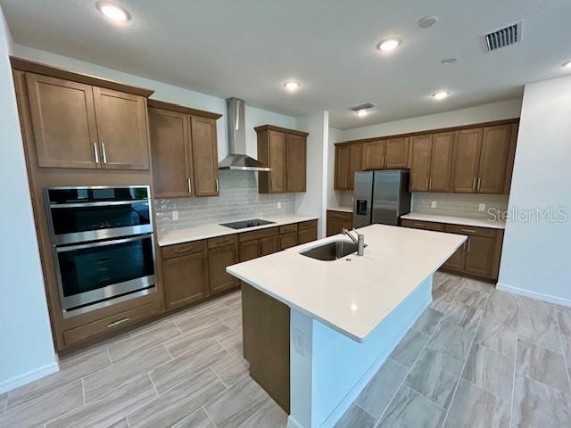 kitchen featuring a center island with sink, wall chimney range hood, backsplash, appliances with stainless steel finishes, and sink