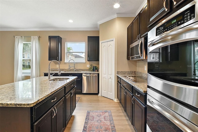 kitchen with a kitchen island with sink, stainless steel appliances, ornamental molding, light stone counters, and sink