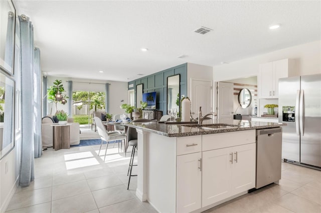 kitchen featuring appliances with stainless steel finishes, dark stone counters, sink, white cabinets, and an island with sink