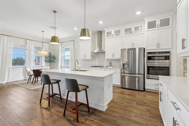 kitchen featuring a center island with sink, stainless steel appliances, hanging light fixtures, wall chimney range hood, and white cabinets