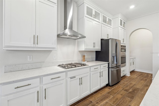 kitchen featuring white cabinets, appliances with stainless steel finishes, wall chimney exhaust hood, ornamental molding, and light stone counters