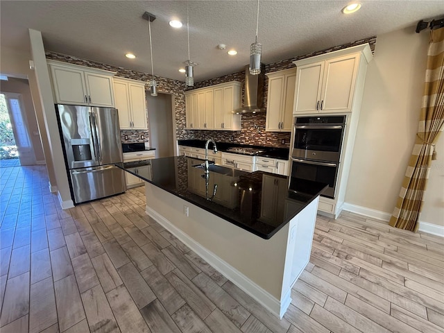 kitchen with appliances with stainless steel finishes, pendant lighting, wall chimney range hood, and a kitchen island with sink