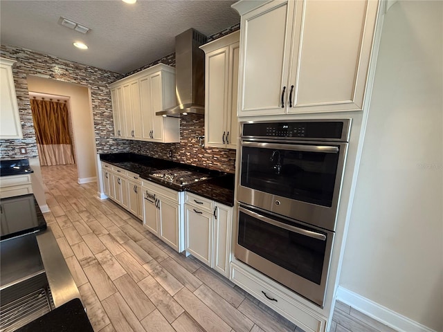 kitchen featuring wall chimney exhaust hood, light hardwood / wood-style flooring, decorative backsplash, dark stone counters, and appliances with stainless steel finishes