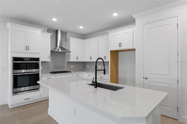 kitchen featuring a kitchen island with sink, wall chimney range hood, sink, white cabinetry, and stainless steel appliances