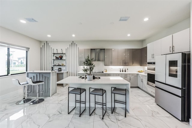 kitchen with appliances with stainless steel finishes, wall chimney range hood, gray cabinets, a breakfast bar area, and an island with sink