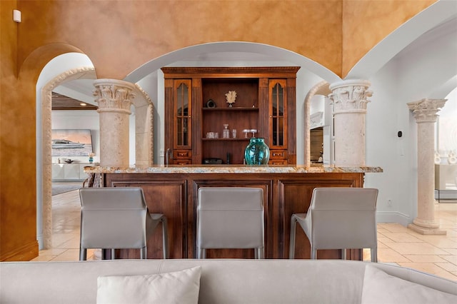 bar featuring a towering ceiling, ornate columns, baseboards, and stone tile flooring