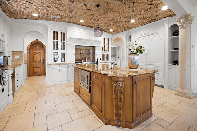 kitchen with brick ceiling, an island with sink, stainless steel microwave, and glass insert cabinets