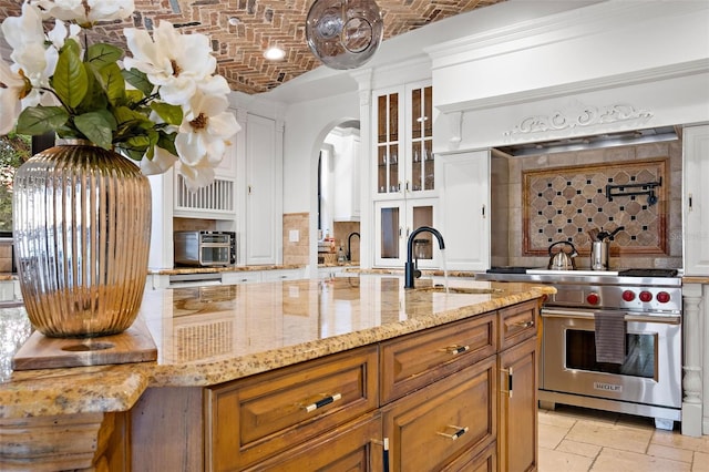 kitchen with arched walkways, glass insert cabinets, premium range, an island with sink, and brick ceiling