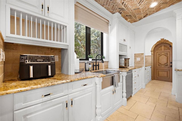 kitchen featuring stone tile flooring, stainless steel dishwasher, white cabinetry, a sink, and brick ceiling