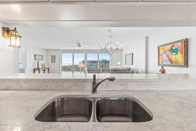 kitchen with light stone counters, sink, pendant lighting, and ceiling fan with notable chandelier