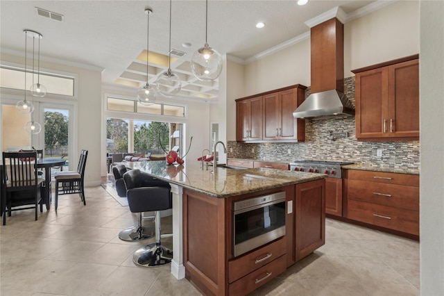 kitchen featuring stainless steel appliances, backsplash, stone countertops, wall chimney range hood, and an island with sink