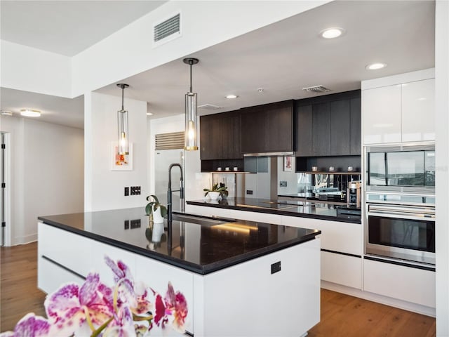 kitchen featuring built in appliances, hanging light fixtures, light hardwood / wood-style floors, and an island with sink