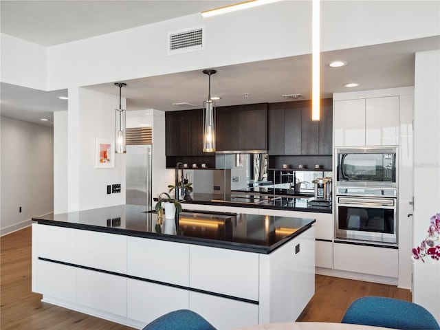 kitchen with pendant lighting, built in appliances, dark brown cabinetry, wood-type flooring, and an island with sink