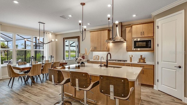 kitchen with black microwave, wall chimney range hood, crown molding, decorative light fixtures, and a kitchen island with sink