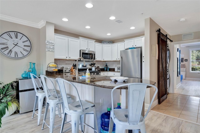 kitchen featuring white cabinets, stainless steel appliances, dark stone countertops, kitchen peninsula, and a barn door