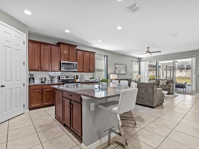 kitchen featuring stainless steel appliances, an island with sink, a breakfast bar, dark stone countertops, and ceiling fan