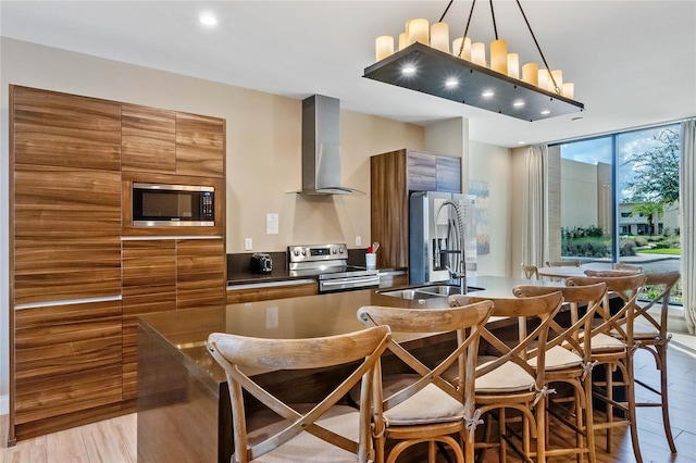 kitchen featuring stainless steel appliances, decorative light fixtures, light wood-type flooring, a wall of windows, and wall chimney range hood