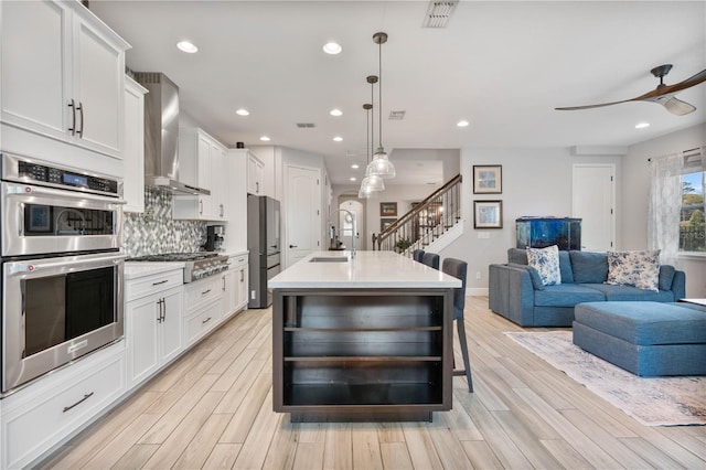 kitchen featuring a kitchen island with sink, appliances with stainless steel finishes, pendant lighting, white cabinets, and wall chimney range hood