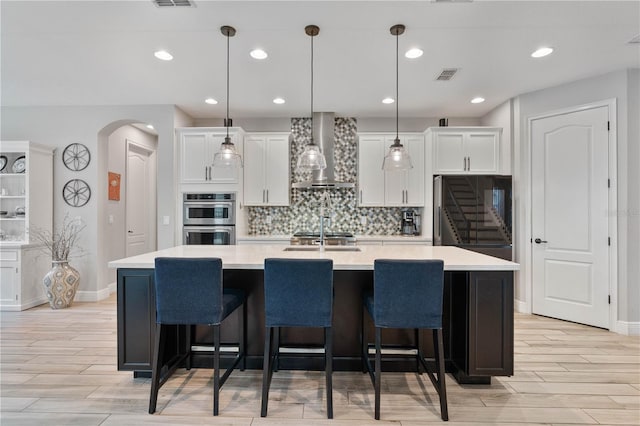 kitchen featuring double oven, wall chimney range hood, a kitchen island with sink, and white cabinetry