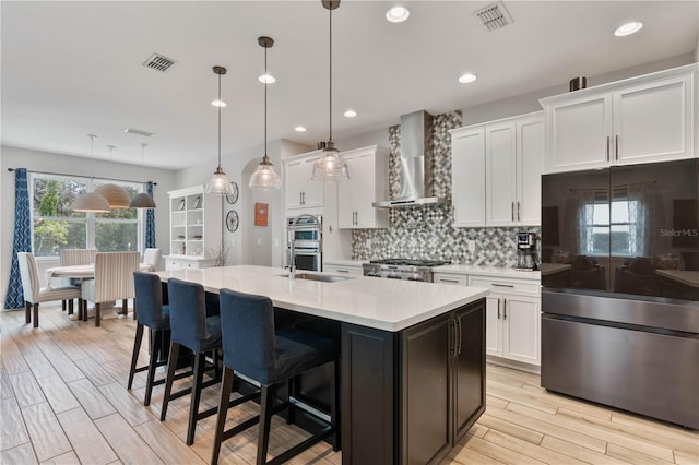 kitchen featuring stainless steel appliances, white cabinets, an island with sink, wall chimney range hood, and pendant lighting