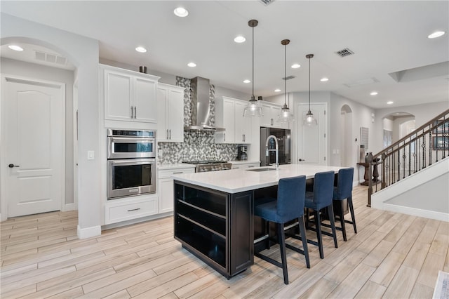 kitchen with stainless steel appliances, decorative light fixtures, white cabinetry, an island with sink, and wall chimney range hood