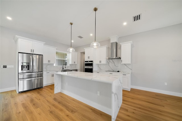 kitchen with a center island, wall chimney exhaust hood, white cabinetry, stainless steel appliances, and hanging light fixtures