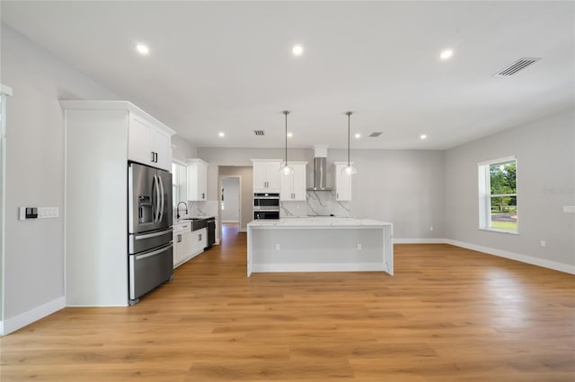 kitchen with appliances with stainless steel finishes, decorative light fixtures, white cabinetry, wall chimney range hood, and light wood-type flooring