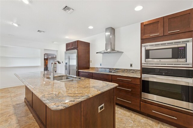 kitchen featuring wall chimney exhaust hood, sink, light stone counters, appliances with stainless steel finishes, and a kitchen island with sink