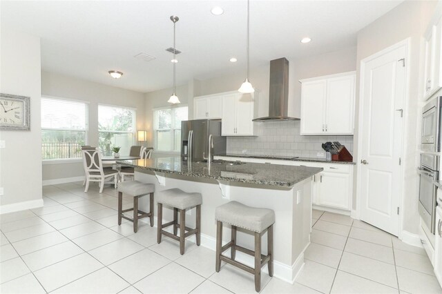 kitchen featuring white cabinetry, dark stone counters, wall chimney exhaust hood, tasteful backsplash, and a center island with sink