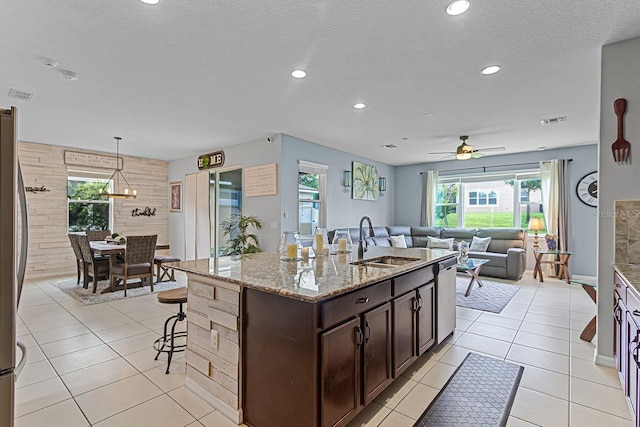 kitchen featuring decorative light fixtures, sink, dark brown cabinetry, light stone counters, and a center island with sink