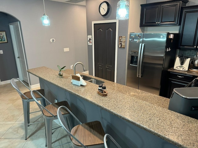 kitchen featuring sink, hanging light fixtures, light stone countertops, stainless steel fridge with ice dispenser, and a breakfast bar area