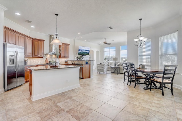 kitchen featuring stainless steel fridge with ice dispenser, wall chimney range hood, ceiling fan with notable chandelier, light stone counters, and a center island