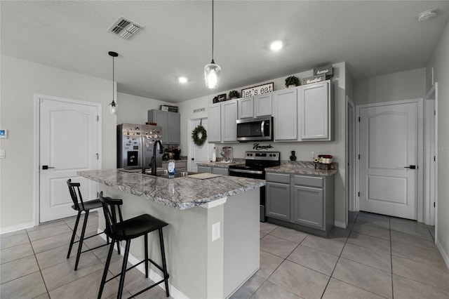 kitchen with a kitchen island with sink, gray cabinets, appliances with stainless steel finishes, hanging light fixtures, and light tile patterned flooring