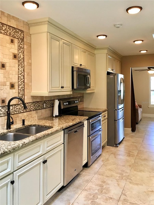 kitchen with appliances with stainless steel finishes, sink, backsplash, cream cabinets, and light stone countertops