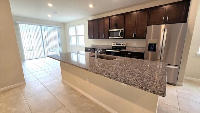 kitchen featuring sink, light tile patterned flooring, a kitchen island with sink, stainless steel appliances, and dark stone counters