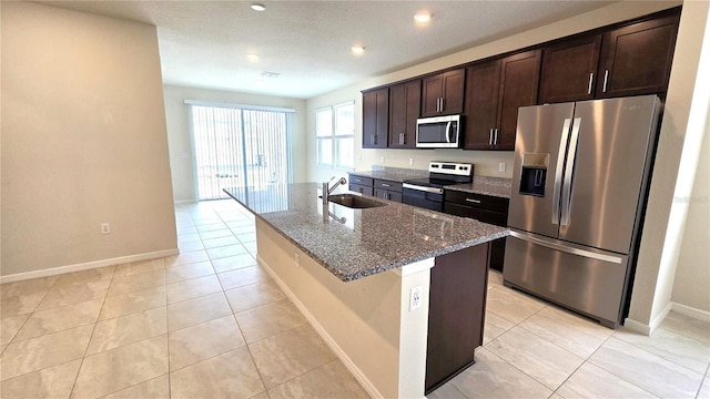 kitchen with dark brown cabinetry, appliances with stainless steel finishes, dark stone counters, sink, and a center island with sink