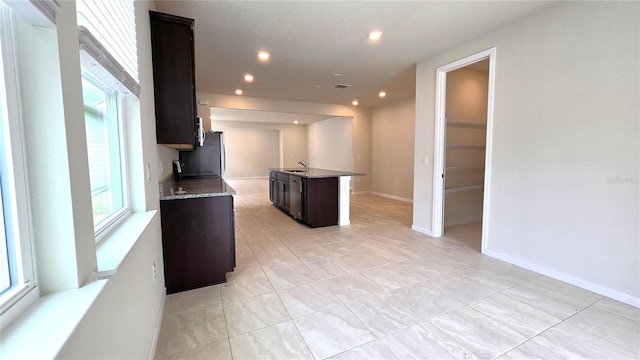 kitchen featuring sink, dark brown cabinets, stainless steel fridge, a center island with sink, and stone counters