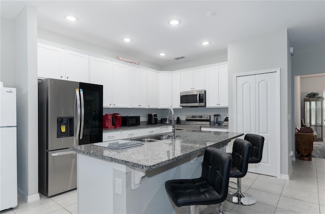 kitchen featuring a center island with sink, sink, white cabinetry, stainless steel appliances, and dark stone counters