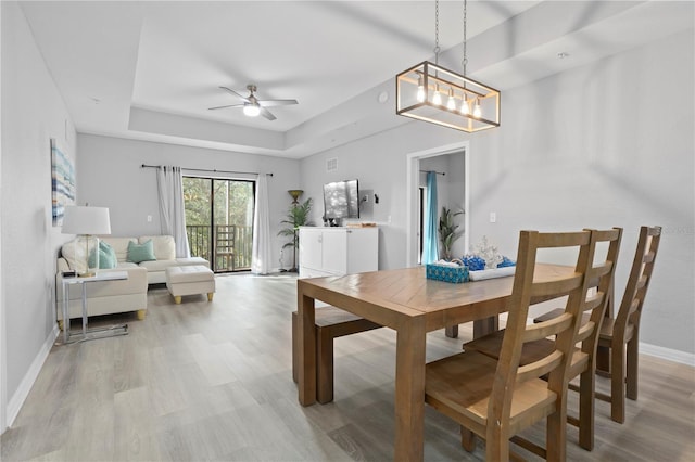 dining space with ceiling fan with notable chandelier, a tray ceiling, and light hardwood / wood-style flooring