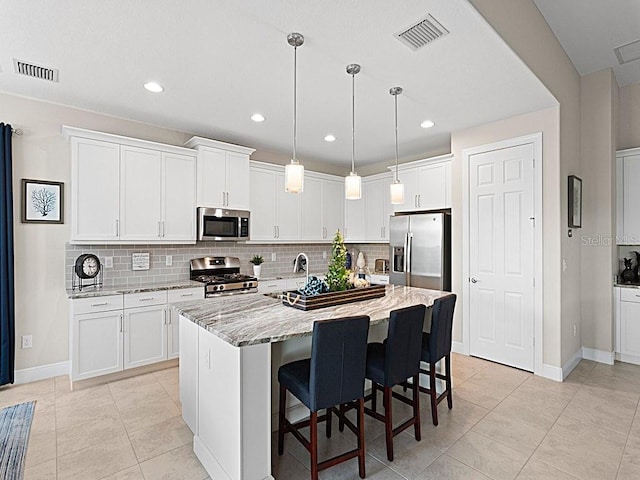 kitchen featuring hanging light fixtures, stainless steel appliances, light stone countertops, an island with sink, and white cabinets