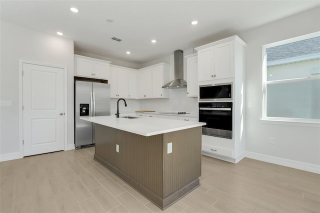 kitchen with appliances with stainless steel finishes, wall chimney range hood, a center island with sink, and white cabinetry