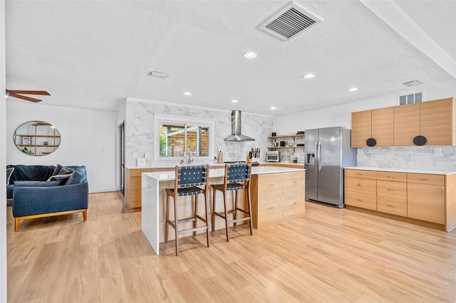 kitchen with a center island, light wood-type flooring, stainless steel refrigerator with ice dispenser, wall chimney range hood, and a breakfast bar