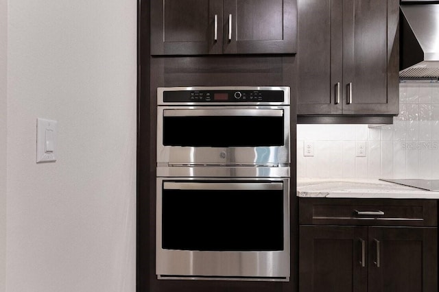 kitchen featuring double oven, tasteful backsplash, light stone counters, dark brown cabinetry, and wall chimney exhaust hood