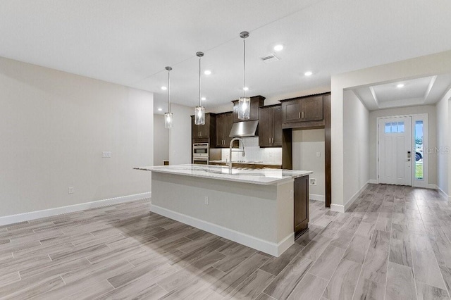 kitchen featuring dark brown cabinetry, light stone counters, light hardwood / wood-style flooring, an island with sink, and pendant lighting