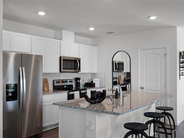 kitchen featuring stainless steel appliances, an island with sink, light stone counters, white cabinets, and tasteful backsplash