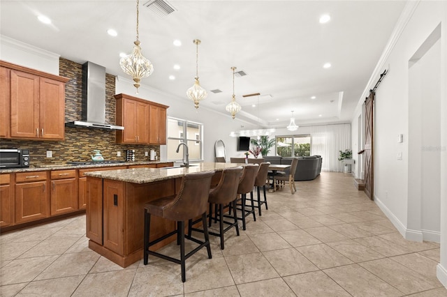 kitchen with a barn door, wall chimney range hood, pendant lighting, a kitchen island with sink, and light stone counters