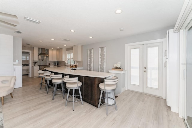 kitchen with wall chimney range hood, light hardwood / wood-style floors, a kitchen bar, sink, and french doors