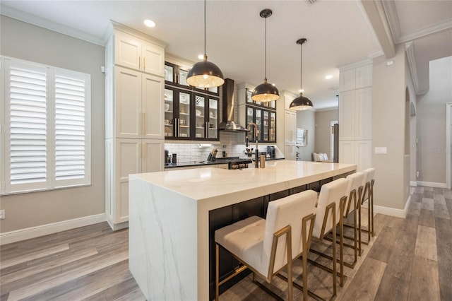 kitchen with hardwood / wood-style flooring, hanging light fixtures, an island with sink, white cabinets, and wall chimney exhaust hood